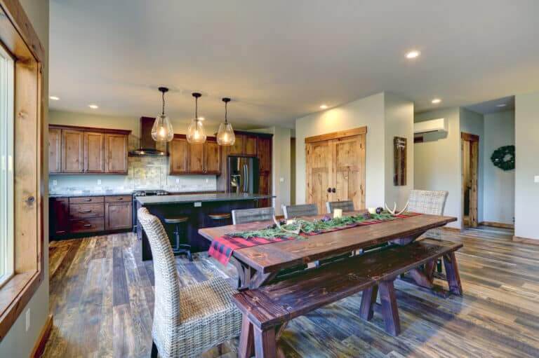 A spacious open-plan kitchen and dining area featuring rustic wooden floors, cabinets, and furniture. The room is warmly lit with pendant lights over the kitchen island and a large dining table adorned with a holiday centerpiece. The wood flooring showcases the craftsmanship of Mr. Sander.