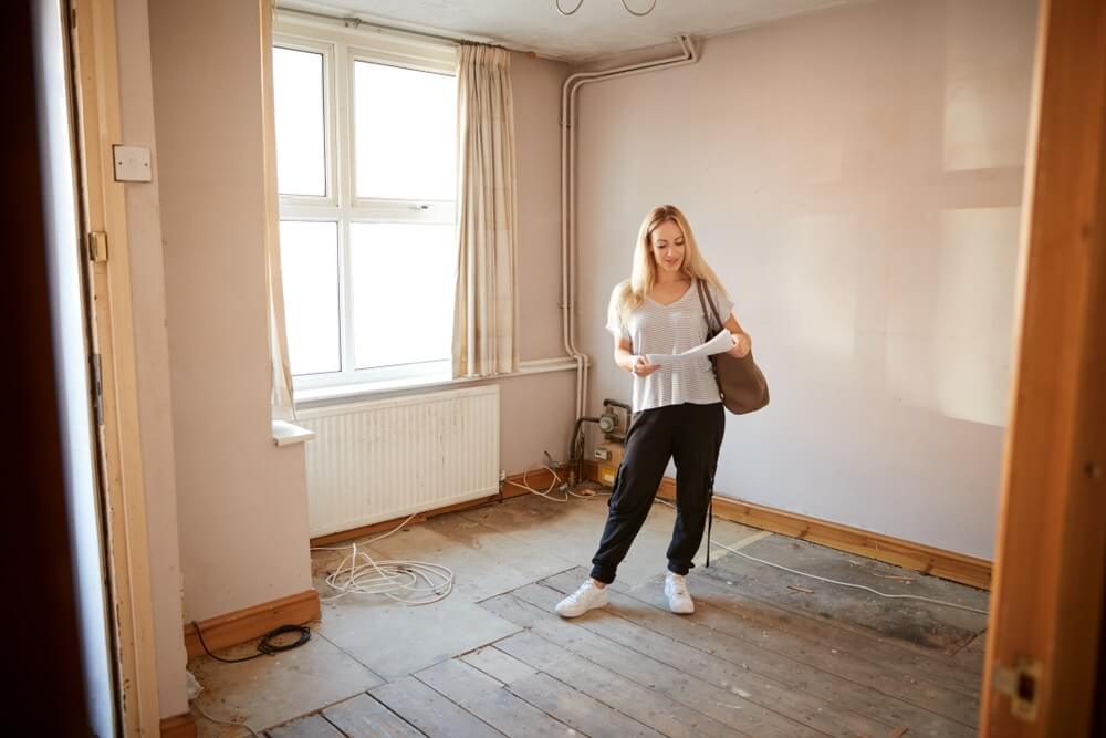A woman stands in a room with bare wooden floors, holding a paper, indicating a need for floor sanding and refinishing. The room has old curtains, exposed wires, and unpainted walls.