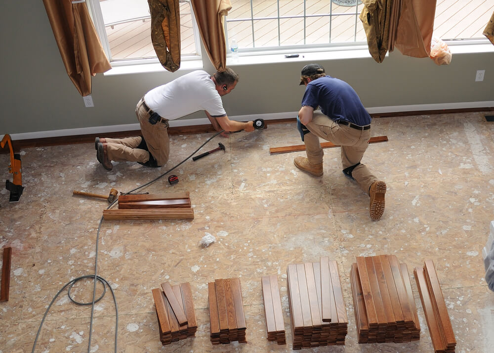 Two professionals from Mr. Sander are installing hardwood flooring in a bright room with large windows. They are meticulously aligning and securing the wooden planks, ensuring a seamless and durable installation. Tools and stacks of wood planks are organized around them for easy access.