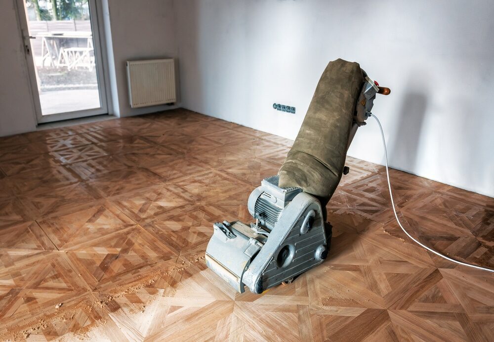 A floor sanding machine on a parquet floor in an empty room.