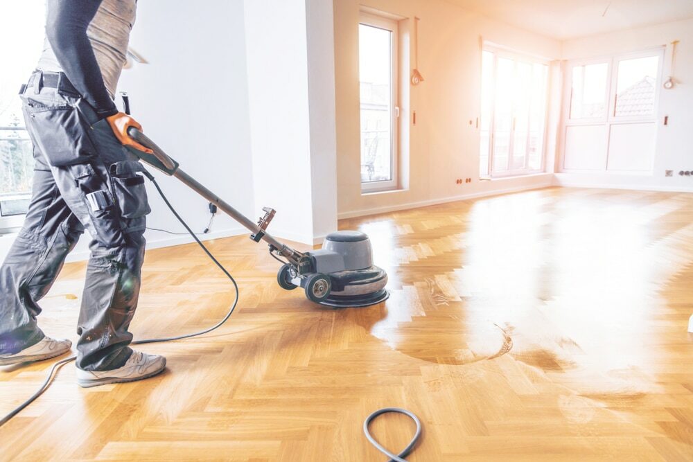 Worker operating a floor polishing machine on a bright wooden floor in a sunlit room.