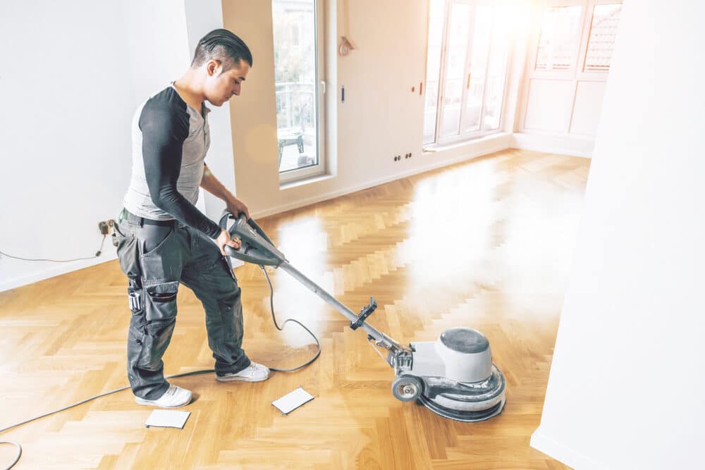 A man operating a professional floor polishing machine on a herringbone-patterned wooden floor in a bright room with large windows.