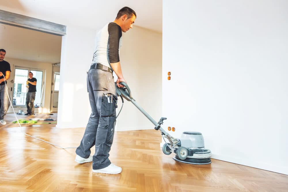 A professional worker using a floor polishing machine on a herringbone parquet floor in a bright, empty room, with another worker in the background.