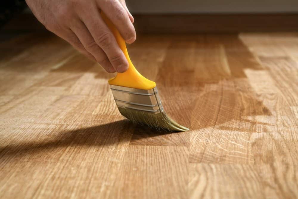 Hand applying finish to wooden flooring with a yellow paintbrush.