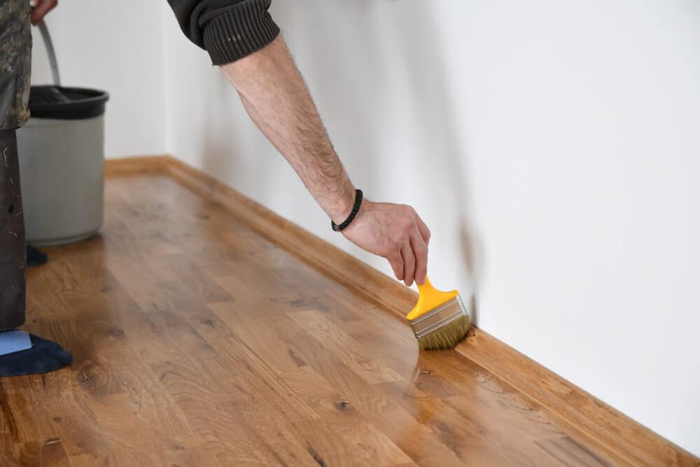 Close-up of a hand applying stain to the edges of a hardwood floor with a brush.