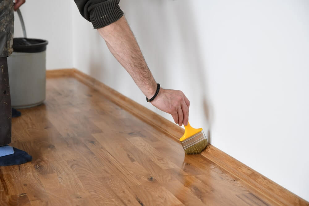 A person applying a finish to an oak wood floor using a brush. The floor has been recently sanded and is being refinished, with the worker carefully coating the edges near the wall.