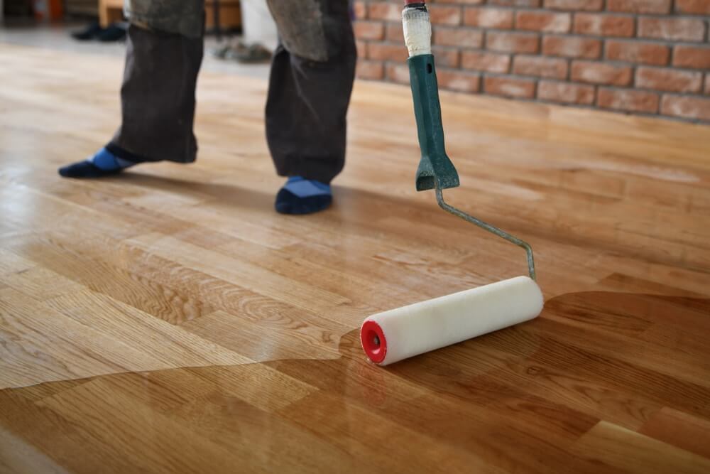 Worker applying finish on a hardwood floor with a paint roller, wearing blue socks, and standing on the floor.