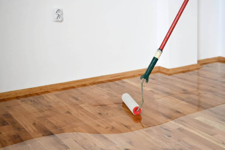 A roller being used to apply a clear finish to a newly sanded wooden floor in a room. The floor has a glossy sheen, reflecting the even application of the finish. An electrical outlet is visible on the wall in the background.