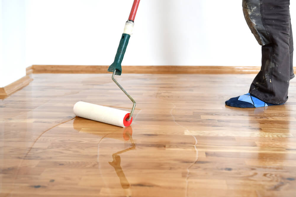 A person applies a glossy finish to a wooden floor using a roller with an extended handle. The floor has a smooth and shiny appearance where the finish has been applied, contrasting with the uncoated area. The person, dressed in work pants and blue protective shoe covers, is partially visible, focused on evenly spreading the lacquer.