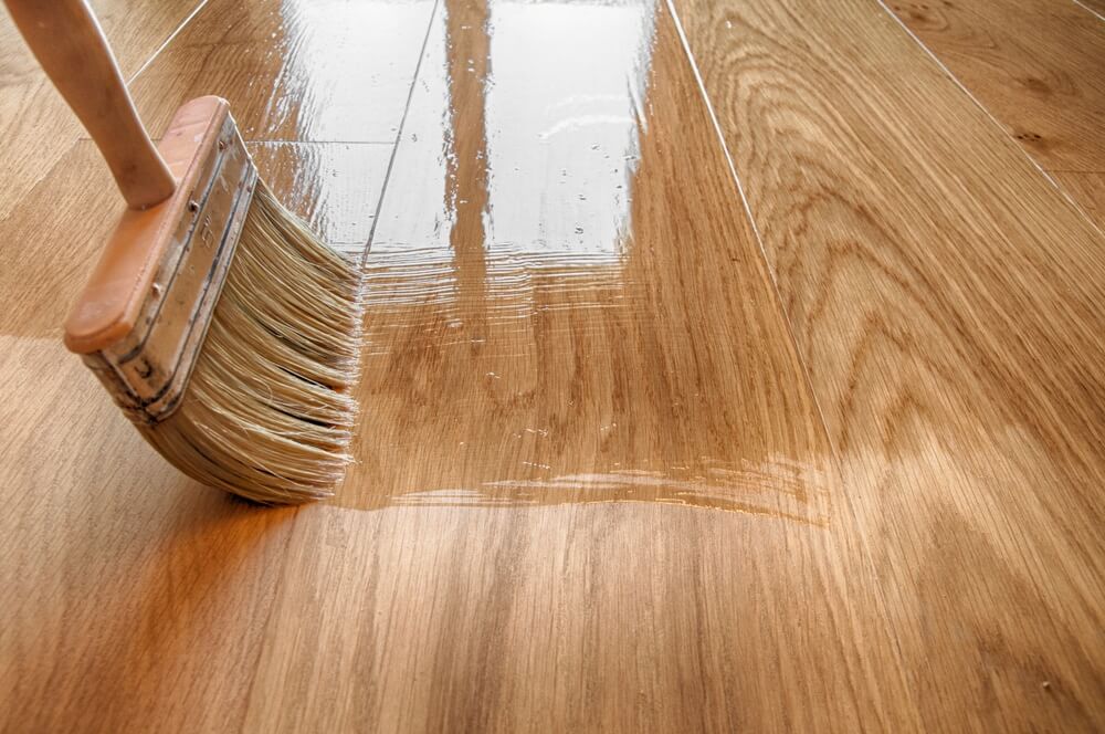Close-up of a paintbrush applying finish on a sanded hardwood floor.