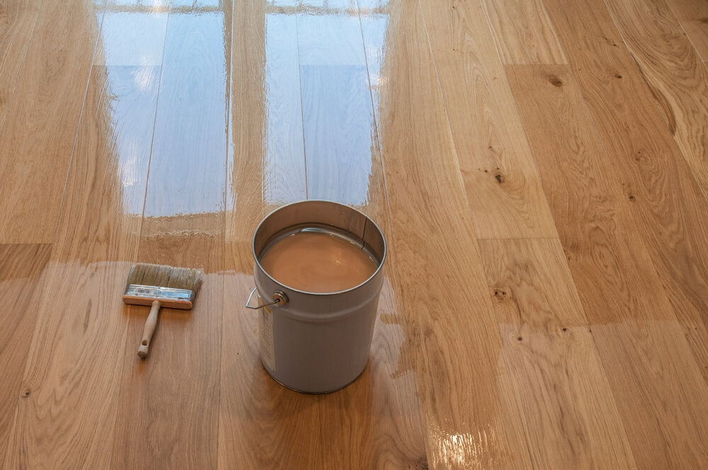 Application of finish on a sanded hardwood floor with a paintbrush and a bucket of finish.