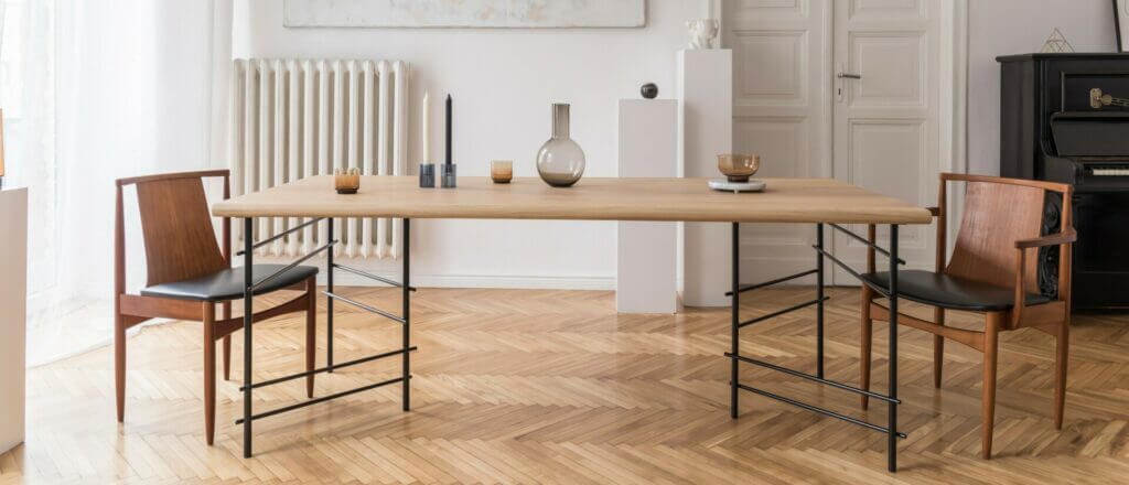 A stylish living room with a newly sanded and refinished herringbone parquet floor by Mr Sander®. The room features a wooden table with two modern wooden chairs, a radiator, and a black piano. The floor's smooth and glossy finish enhances the room's elegant ambiance.