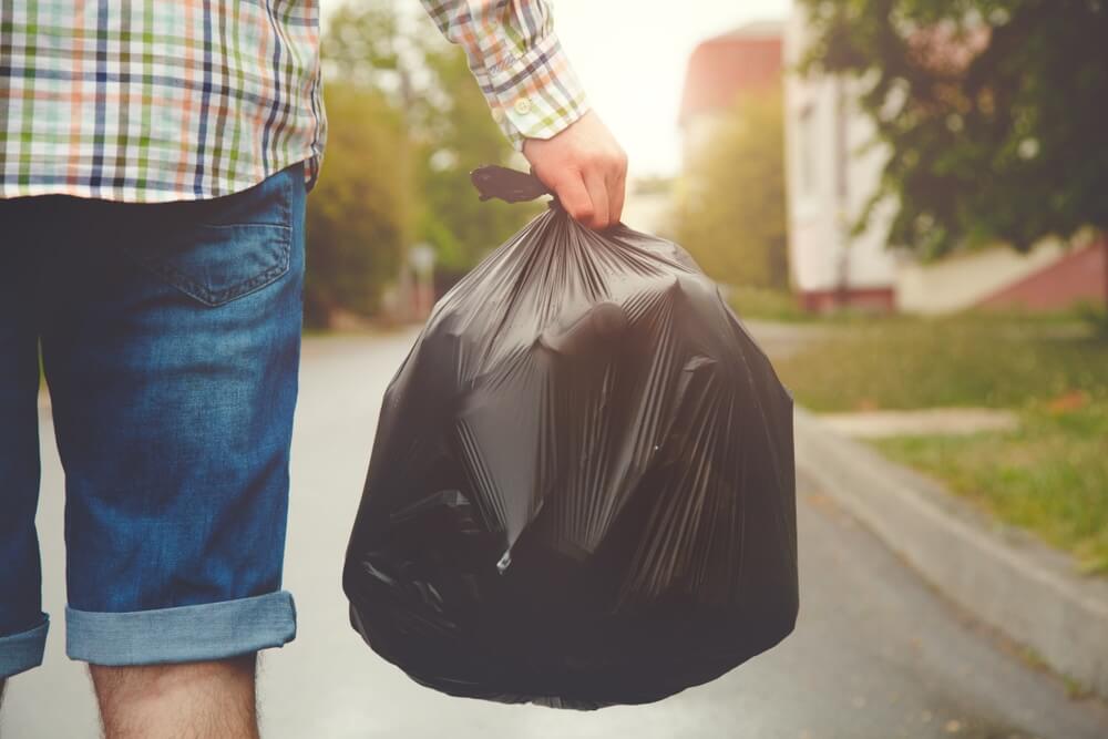 A person in casual attire, wearing a checkered shirt and denim shorts, carries a black garbage bag outdoors. The scene signifies cleanup efforts, possibly after a floor sanding and refinishing project.