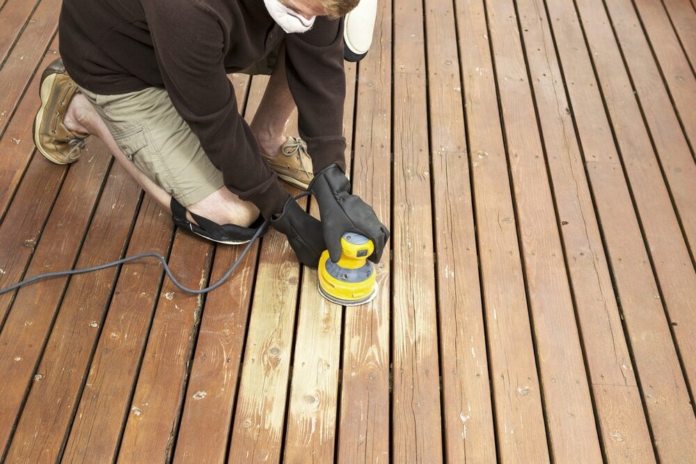 Person kneeling while using an electric sander on a wooden deck.