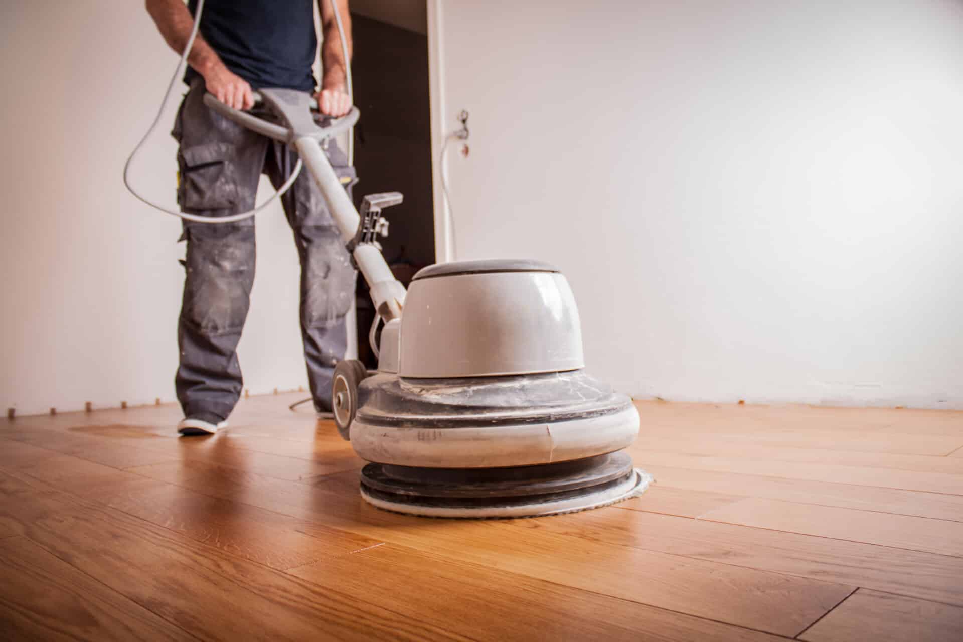 Person operating a floor buffer on a wooden floor, seen from a low angle.