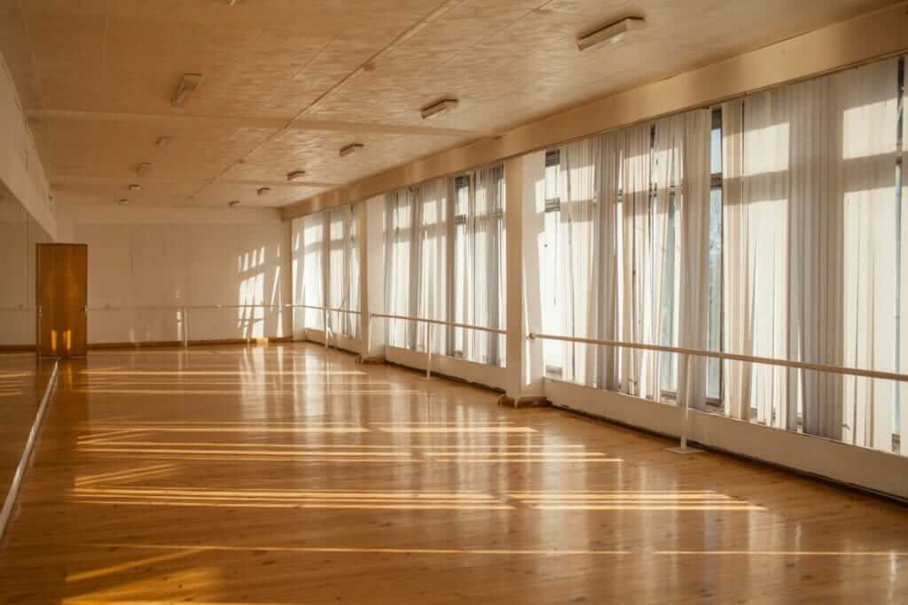 A spacious dance studio with a beautifully refinished pine wood floor, polished to a high gloss. The room is flooded with natural light from large windows covered with sheer curtains, casting shadows across the floor. The open space features ballet barres along the windows and clean white walls, providing a bright and airy environment. The smooth and glossy finish of the pine floor enhances the room's elegance and functionality for dance or exercise activities.