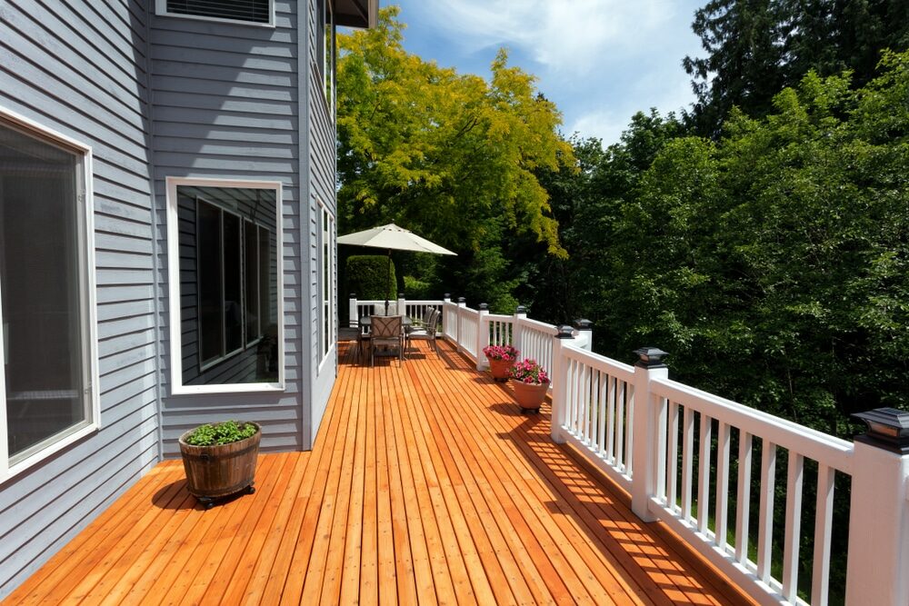 Sunny deck with a wooden floor and white railings attached to a grey house.