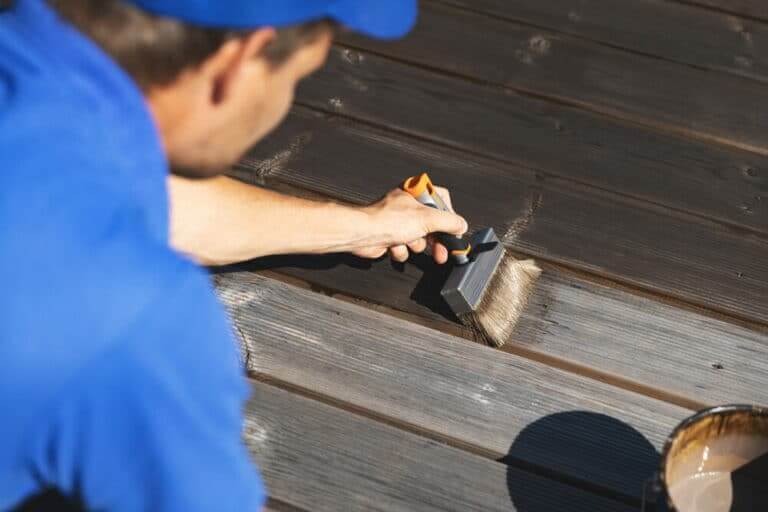 Person in a blue shirt applying dark stain to wooden decking with a brush.