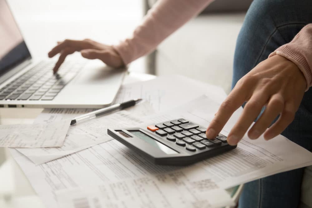 Close-up of a person using a calculator and laptop amidst scattered papers on a desk.