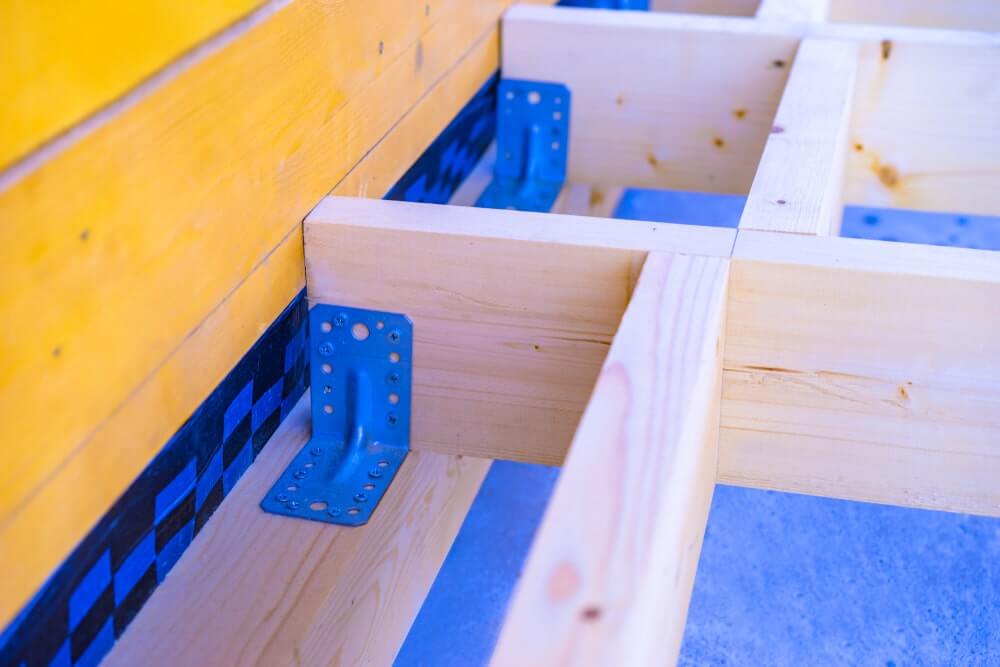 A close-up view of a wooden floor frame under construction, featuring light pine wood beams securely fastened with blue metal brackets. The structure is being prepared for flooring installation or refinishing.