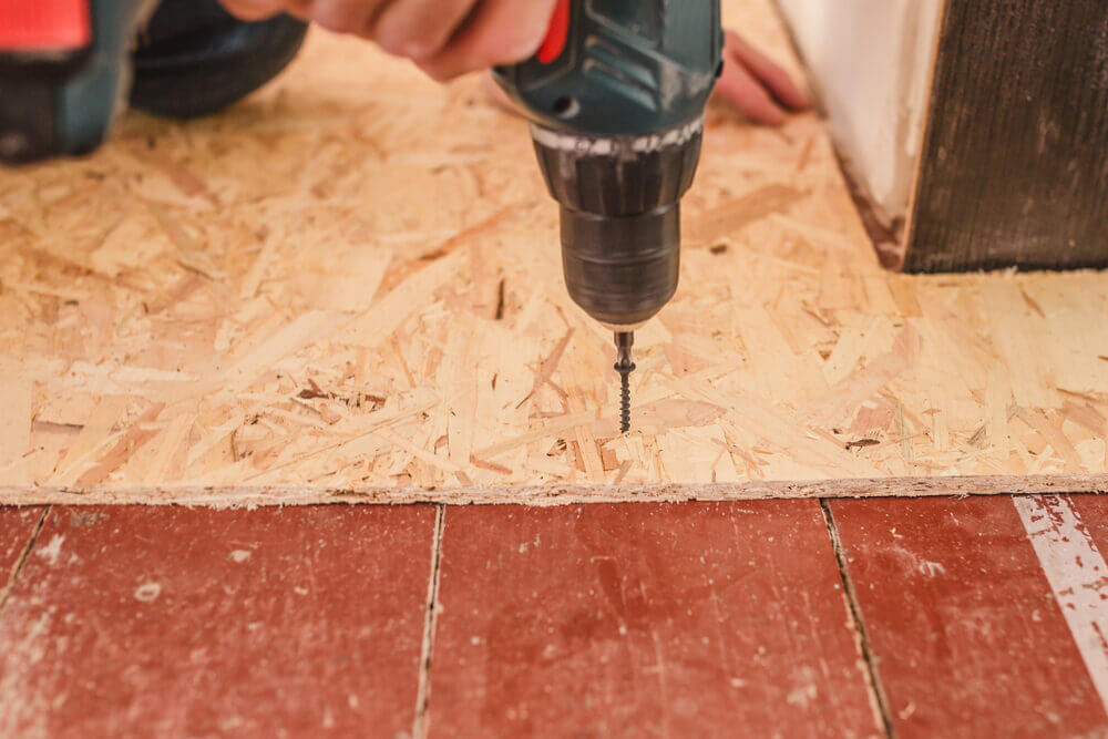 A close-up of a worker using a power drill to secure an OSB board over an older wooden floor during subfloor preparation.