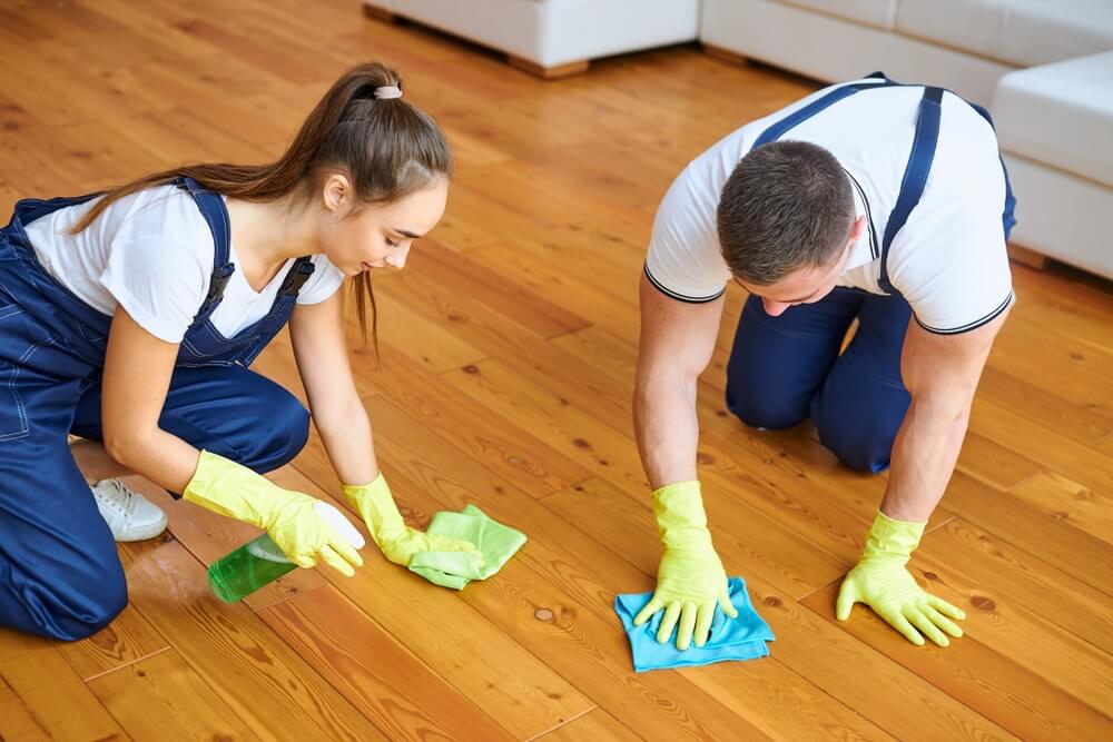 Two professional cleaners wearing gloves and overalls, scrubbing and polishing a hardwood floor to a pristine finish, showcasing their dedication to cleanliness and detail.