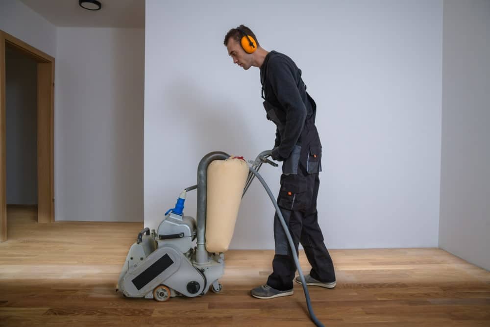 A man from Mr Sander® is sanding a floor in a UK living room using a Bona Belt sander (model AMO110000.3). This professional machine features a 2.2 kW motor, 230 V, 50 Hz, and a drum width of 200 & 250 mm. The sanding belt size is 200 x 750 mm or 250 x 750 mm, with a total weight of 74 kg. The room has blue columns, green hanging plants, and a bar area with green tiles.