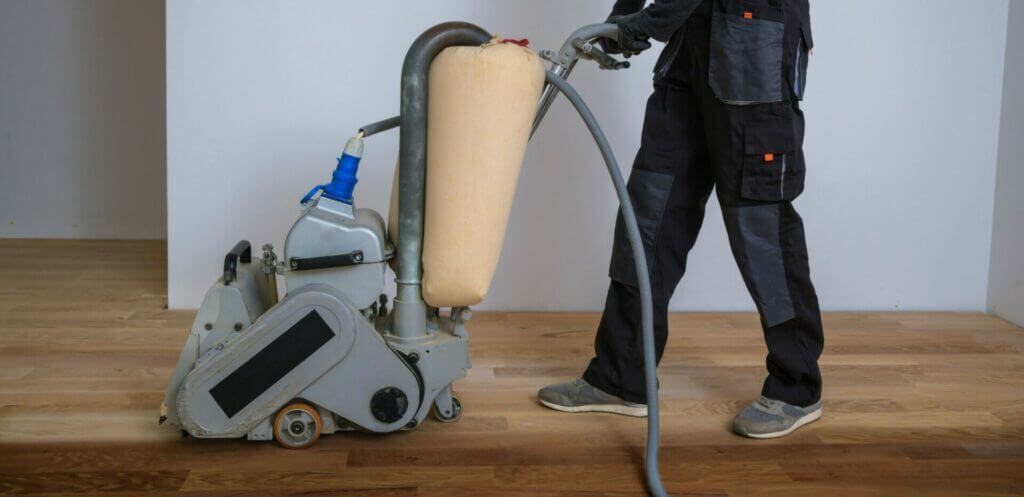 A professional from Mr Sander® sands a living room floor using a Bona Belt (AMO110000.3) belt sander. The machine has a motor power of 2.2 kW, voltage of 230 V, frequency of 50 Hz, drum width of 200 & 250 mm, and a sanding belt size of 200 x 750 mm or 250 x 750 mm. The total weight of the machine is 74 kg.