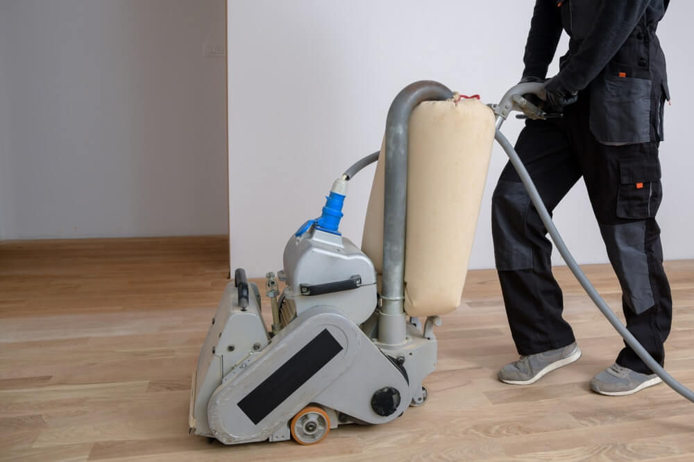 A professional from Mr Sander® using a Bona Belt sander (model AMO110000.3) to sand a floor in a living area in the UK. The belt sander has an effect of 2.2 kW, operates at 230 V and 50 Hz, and measures 250x750 mm. The drum width is 200 & 250 mm, and the sanding belt size is 200 x 750 mm or 250 x 750 mm. The motor power is 2.2 kW, and the total weight of the machine is 74 kg.