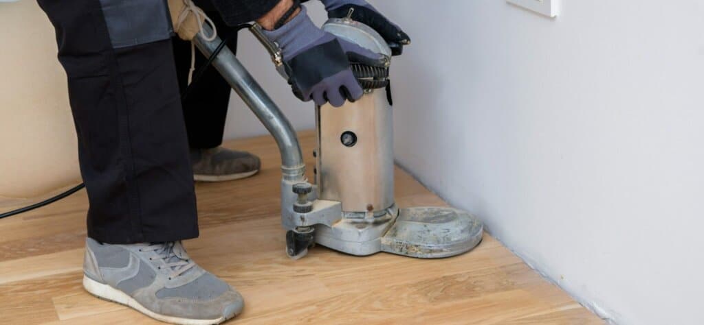 A professional from Mr Sander® using a Bona Edge UX edger (model AMO230600) to sand the floor edges in a living area in the UK. The Bona Edge UX has a motor power of 1.25 kW, operates at 230V-50Hz, with sanding arm lengths of 130 mm and 220 mm, and a disc size of 178 mm. The edger weighs 14.4 kg with a 130 mm arm and 15.4 kg with a 220 mm arm. It features an LED strip light and operates at a noise level of 92 dB with a disc speed of 3000 rpm.
