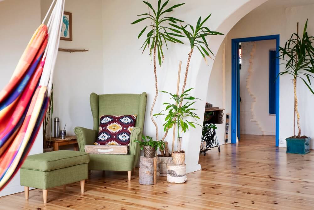 A cozy and vibrant living room in, UK, featuring a green armchair with a colorful patterned cushion, a matching ottoman, and several potted plants. The room is decorated with a colorful hammock, white arched doorways, and blue door frames, highlighting a cheerful and eclectic interior design with polished wooden floors.