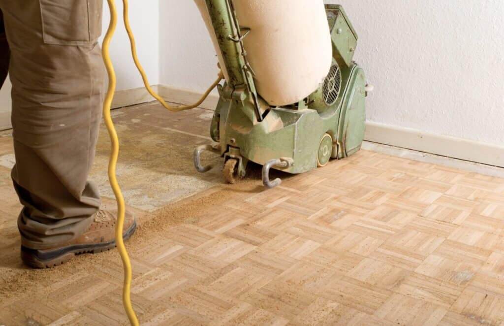 A Mr Sander® technician sanding a parquet floor in a East Meon, UK living room using a LAGLER HUMMEL® machine. Features include powerful LED lighting, high powered aggressive sanding, extremely low dust emissions, quick belt changes, 30+ years lifespan, and easy maintenance. Technical specs: 230 V / 50 CPS / 2.2 kW motor, 200 mm drum width, 2400 rpm drum speed, 200 x 750 mm belt dimensions, 79 kg weight.