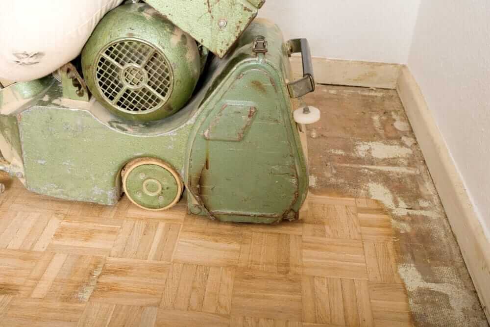 A man from Mr Sander® is sanding a parquet floor in a living room in Finsbury Park, UK, using the Lagler HUMMEL®. The machine features a powerful LED lighting system, high-powered aggressive sanding, low dust emissions, easy belt changes, and a 79 kg single-phase AC motor with thermal overload protection.