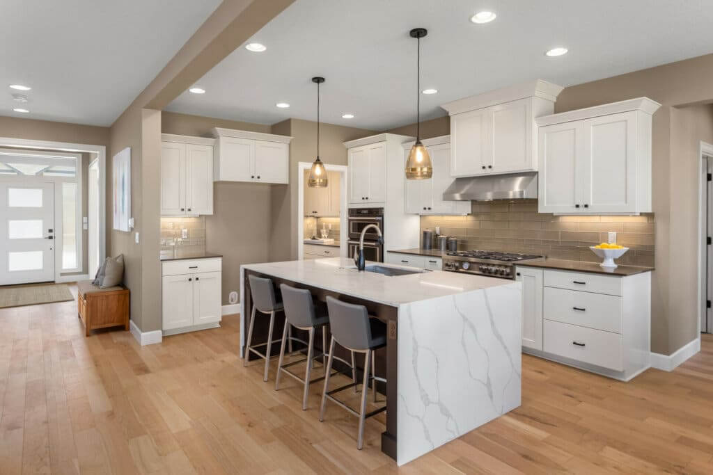 A modern kitchen with white cabinetry, stainless steel appliances, and a large marble island with seating for three. The kitchen is well-lit with recessed lighting and pendant lights above the island. The floor features light-colored wood, showcasing the sanding and refinishing work done by Mr Sander®.