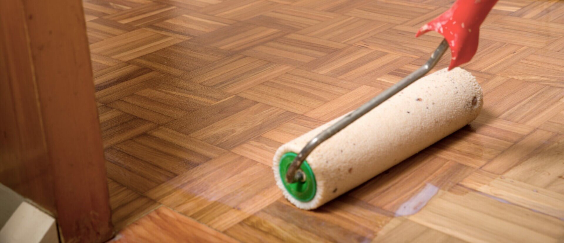 A man from Mr. Sander is applying lacquer to a five-finger parquet wood floor in the living room of a UK home. The detailed parquet pattern is highlighted as the roller brush smoothly spreads the lacquer, enhancing the wood's natural beauty and providing a protective finish.