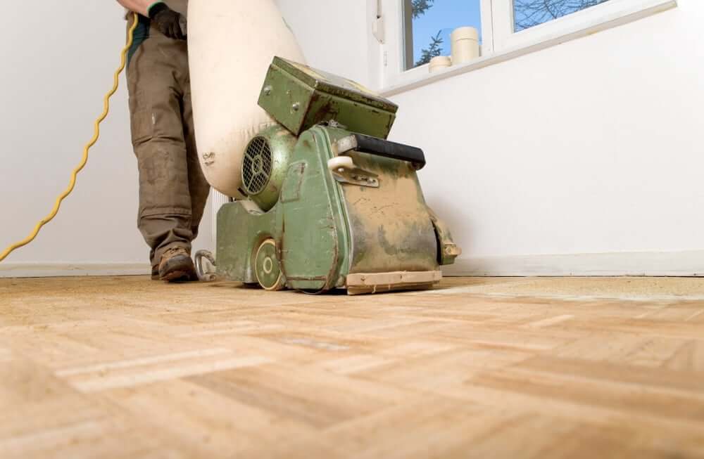 A Mr Sander® technician sanding a parquet floor in a Honor Oak,UK living room using a LAGLER HUMMEL® machine. Features include powerful LED lighting, high powered aggressive sanding, extremely low dust emissions, quick belt changes, 30+ years lifespan, and easy maintenance. Technical specs: 230 V / 50 CPS / 2.2 kW motor, 200 mm drum width, 2400 rpm drum speed, 200 x 750 mm belt dimensions, 79 kg weight.