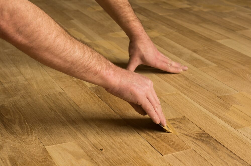 A person's hands repairing a small damaged area on a wooden floor.