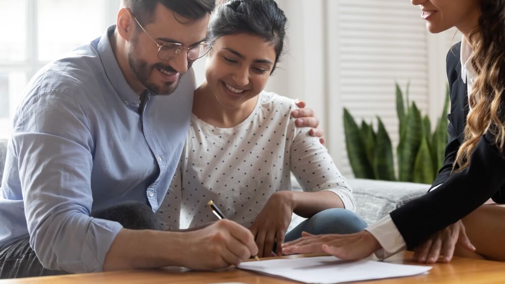  A happy couple signing documents with a professional, sitting in a bright room featuring a beautifully refinished oak hardwood floor by Mr Sander®.