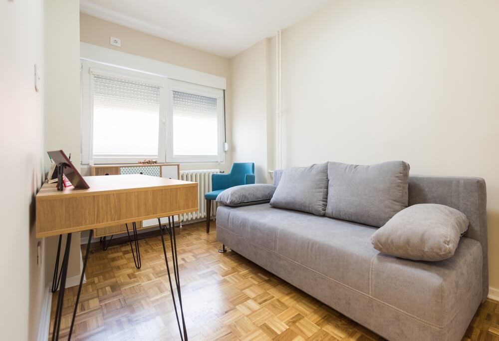 A cozy living room in Stoke Newington, UK, featuring beautifully sanded and refinished parquet flooring done by Mr Sander®. The room includes a gray sofa, a wooden desk, and a blue chair, with large windows letting in natural light. The parquet floor showcases intricate patterns and a smooth finish, highlighting the high-quality restoration work by Mr Sander®.