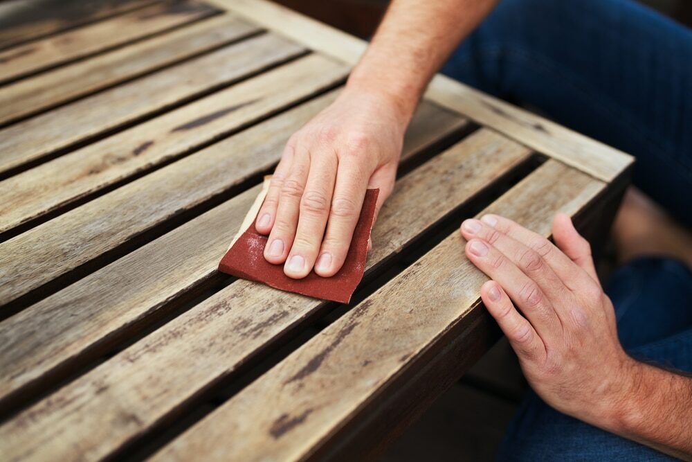 Close-up of a person's hands sanding a wooden bench with sandpaper.