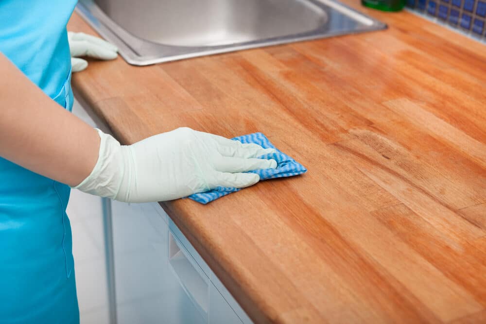 Person in blue apron and gloves cleaning a wooden countertop with a blue cloth.