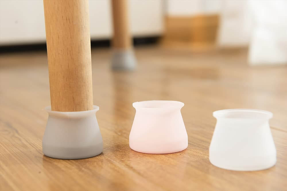 Close-up of furniture legs protected with silicone caps to prevent scratches on a newly refinished wooden floor. The image shows a gray cap fitted on one leg and pink and white caps next to it, ensuring the floor remains in pristine condition.