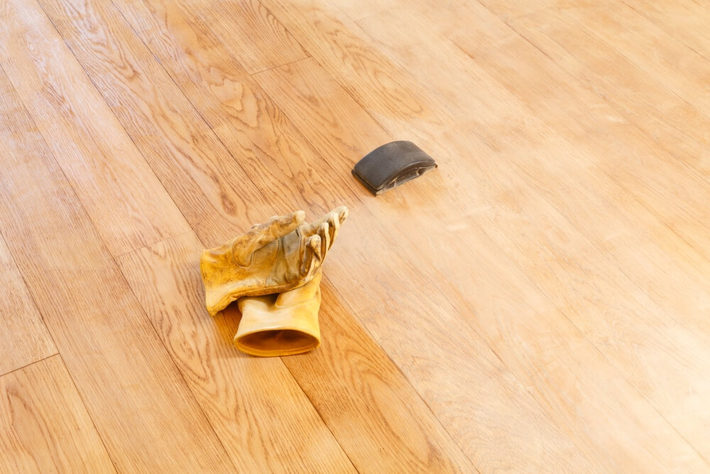 Close-up of a newly sanded wooden floor with a pair of work gloves and a sanding block lying on it, indicating preparation for further refinishing work.