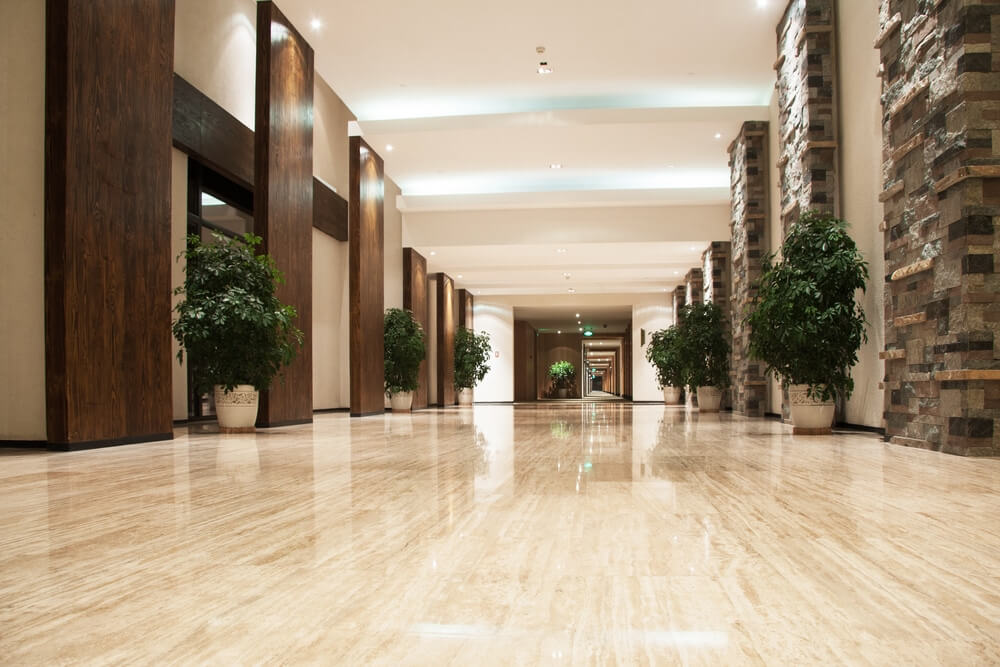 A beautifully polished hallway with gleaming marble floors, showcasing the refinishing work done by Mr Sander®. The hallway features tall wooden columns, stone accent walls, and potted green plants evenly spaced along the sides. Recessed ceiling lights provide bright illumination, highlighting the clean, shiny surface of the floor.