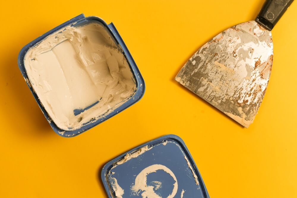 A container of white wood filler and a scraper placed on a yellow surface, used for filling gaps before floor sanding and refinishing.