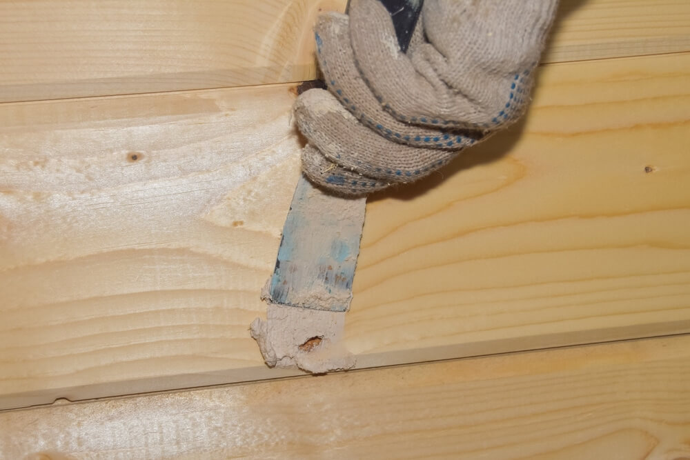 A person wearing a glove applies wood filler to a knot hole in a wooden panel using a putty knife. The wood panels are light-colored and have a natural grain pattern.