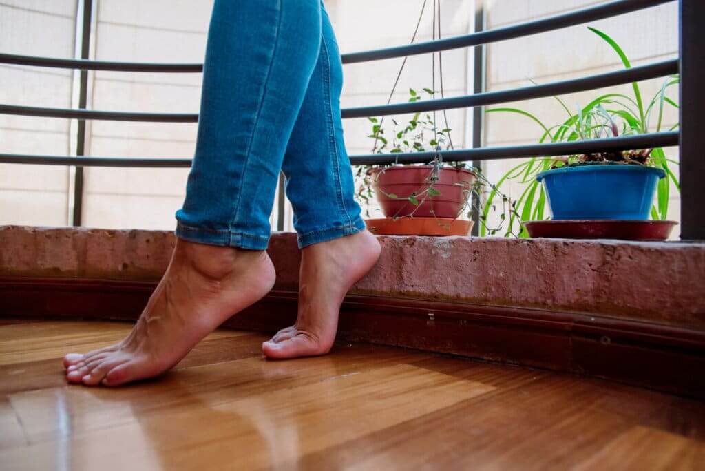 A person standing barefoot on a newly refinished hardwood floor, showcasing its glossy finish and smooth texture. The floor, likely made of oak or maple, is illuminated by natural light coming through a window, with potted plants in the background, highlighting the warm and inviting atmosphere of the space. The refinishing process was completed by Mr. Sander®.
