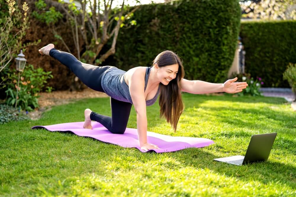 A woman exercises on a yoga mat in her backyard, extending one arm and the opposite leg while smiling at a laptop screen placed on the grass. She appears to be participating in an online workout session.