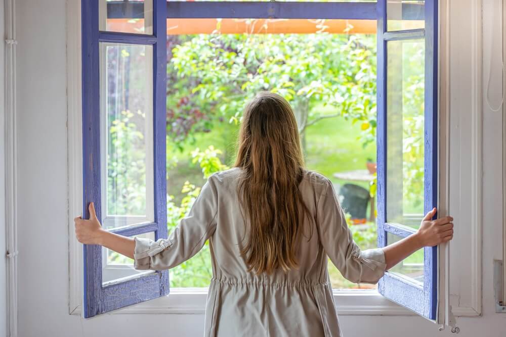 A woman standing by an open window, looking out into a lush, green garden. She holds the window frames, which are painted in a vibrant blue color. Fresh air flows into the room, indicating a well-ventilated space after home renovations such as floor sanding or refinishing.