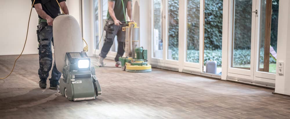 Workers using professional sanding machines on a wooden floor inside a bright room.